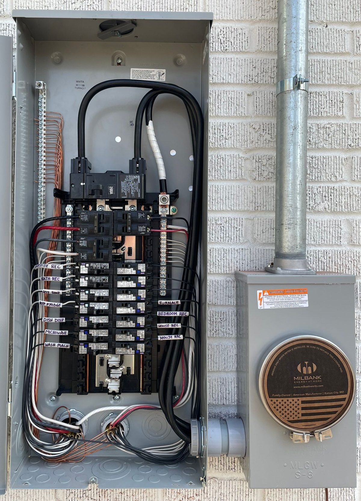 Gray electrical panel on a brick wall with wires and a meter to the right.